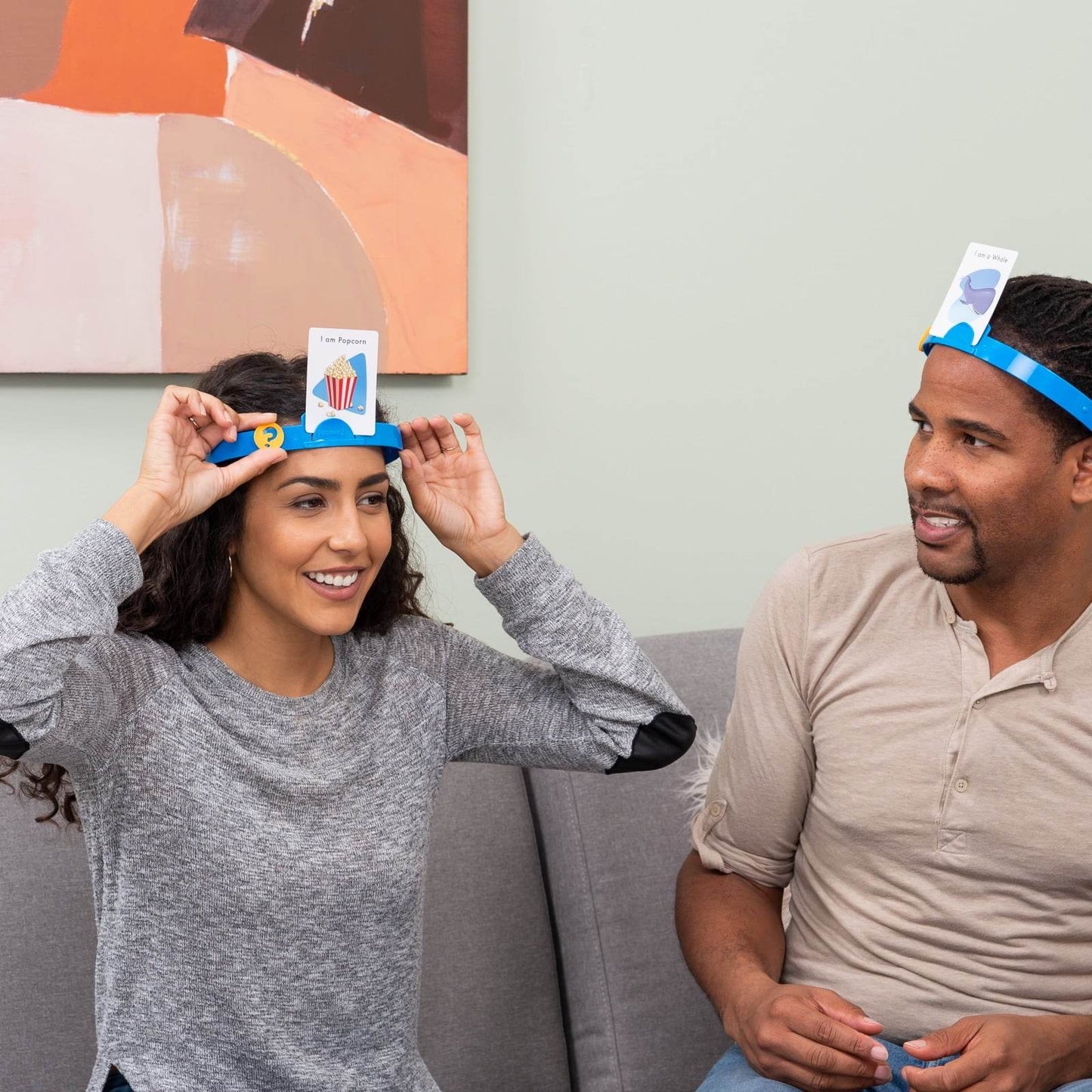 Two adults playing Hedbanz family game India sitting on couch wearing blue headbands with cards