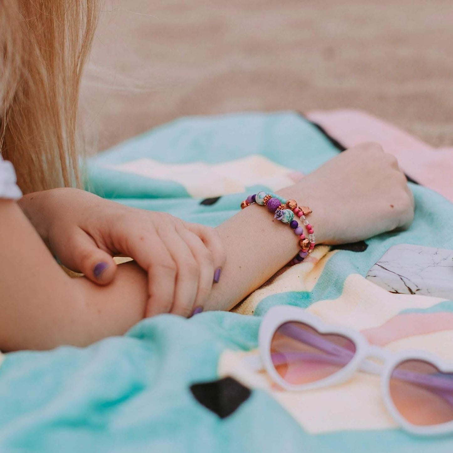 Girl wearing handmade colorful bracelet from DIY jewelry kit for girls lying on a beach towel with heart-shaped sunglasses