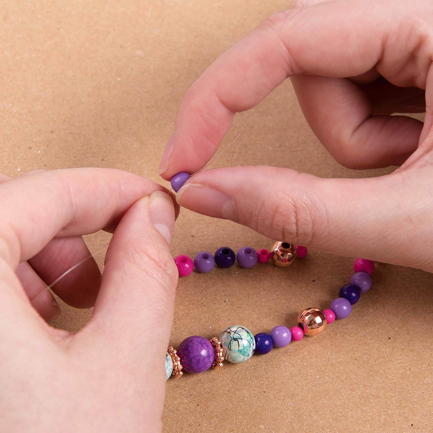 Close-up of hands stringing colorful beads from DIY jewelry kit for girls on a brown surface