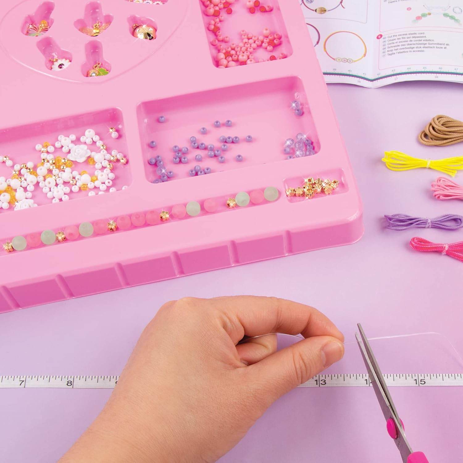 DIY jewelry kit for kids with colorful beads, threads, and a hand cutting thread with scissors on a pink tray