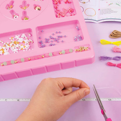 DIY jewelry kit for kids with colorful beads, threads, and a hand cutting thread with scissors on a pink tray